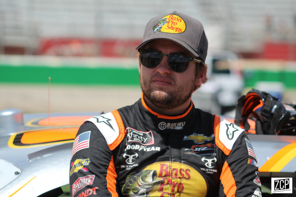 NASCAR driver Noah Gragson leaning against a car in a racing suit